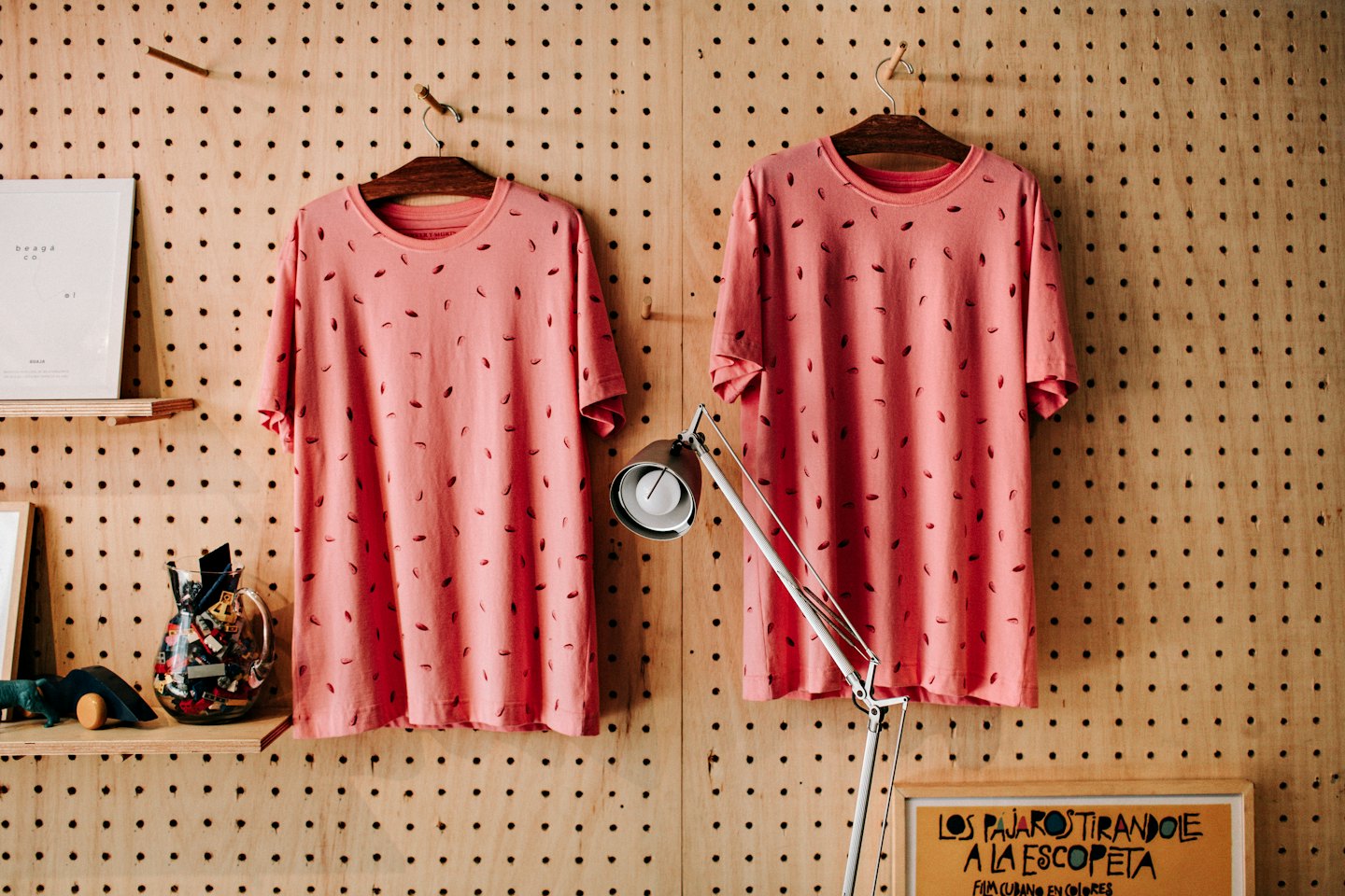 Dwa wzorzyste, koralowe T-shirty wiszą na drewnianej ścianie z pegboardu.